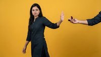 Young Hispanic businesswoman in dark suit making stop gesture refusing handshake on yellow background. Professional rejection concept for workplace communication.