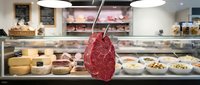 Raw red meat hanging from hook in front of butcher shop display case with products and counter.
