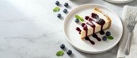 Cheesecake slice with blueberry sauce on white plate, fresh berries and mint garnish, elegant dessert presentation for restaurant menu or food blog.