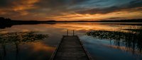 Wooden dock extending into calm lake at golden sunset with dramatic orange sky clouds reflecting in still water surrounded by reeds and natural shoreline.