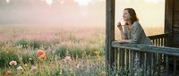 Woman in casual denim shirt leaning on wooden railing of rustic cabin porch, gazing at expansive wildflower meadow at sunset. Serene countryside landscape for wellness and lifestyle content.