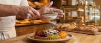 Pastry chef dusting powdered sugar onto fresh fruit tart in bakery kitchen with bread and warm lighting, suitable for culinary, food service, and baking industry content.
