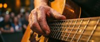 Close-up of elderly musician's weathered hands playing acoustic guitar strings with warm stage lighting creating intimate concert atmosphere.