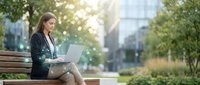 Professional businesswoman working on laptop while sitting on park bench in urban setting with natural sunlight filtering through trees.