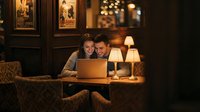 Young Caucasian couple smiling together at laptop in cozy dimly lit restaurant, warm lamp glow, vintage posters, romantic evening date.