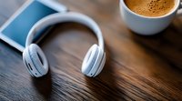 White wireless headphones on dark wooden table with coffee cup and mobile device in background, creating modern lifestyle workspace atmosphere.
