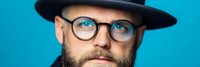 Bearded Caucasian man with glasses and black hat looking directly at camera against bright blue background for portrait photography and lifestyle content.