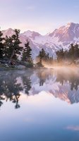 Serene mountain lake reflects snow-capped peaks at sunrise with evergreen trees and misty atmosphere creating peaceful wilderness landscape.