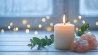 Lit white candle with eucalyptus branch and pink salt crystals on wooden surface, warm bokeh lights creating cozy spa atmosphere for relaxation.