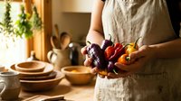 Woman in linen apron holding fresh colorful bell peppers in rustic kitchen with wooden bowls and hanging herbs creating warm cooking atmosphere.