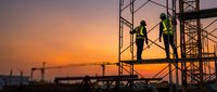 Construction workers in safety vests on scaffolding at sunset. Industrial building site with crane silhouettes against orange sky for construction industry projects.