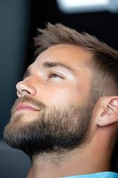 Profile portrait of young Caucasian man with well-groomed beard and modern haircut looking up with serene expression against dark background.