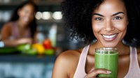 Young African American woman smiling while holding fresh green smoothie in glass, healthy lifestyle concept with natural detox drink and vegetables in background.