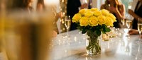 Yellow roses centerpiece in glass vase on elegant dining table with warm ambient lighting and blurred guests in background for wedding reception decor.