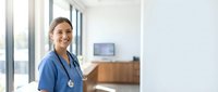 Smiling young Asian female nurse in blue scrubs with stethoscope standing in modern hospital corridor with natural light from windows.