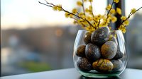 Easter eggs with natural marble effect in glass vase decorated with yellow spring flowers against blurred window background, soft morning light.