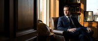 Confident businessman in navy suit sitting in luxury leather chair in elegant office library with warm lighting and wooden bookshelves for corporate success concepts.