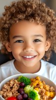 Happy mixed-race child with curly hair smiling while eating healthy colorful meal with quinoa, broccoli, grapes and vegetables for nutrition education.