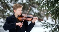 Young Caucasian male violinist performing classical music outdoors in winter snow among evergreen trees, wearing black formal attire for concert or recital.