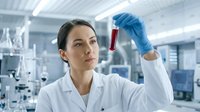 Female scientist in white lab coat and blue gloves examining red liquid sample in test tube within modern laboratory setting, suitable for healthcare, research, and medical education content.