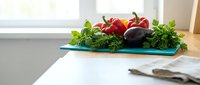 Fresh organic vegetables including red bell peppers, eggplant and herbs arranged on wooden cutting board in bright modern kitchen for healthy cooking preparation.