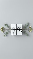 White gift box with black ribbon surrounded by eucalyptus branches and small flowers on neutral background for holiday celebrations and special occasions.