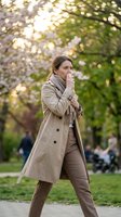 Woman in beige trench coat standing in spring park with blooming trees, wearing casual neutral outfit, outdoor fashion portrait for lifestyle and seasonal content.