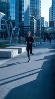 Young male athlete jogging through urban plaza with modern skyscrapers, concrete seating, and blue sculpture installations. Fitness and active lifestyle in contemporary city environment.
