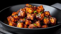 Glazed tofu cubes in sweet and spicy sauce garnished with fresh herbs served on black plate, close up view of Asian vegetarian cuisine with caramelized coating.