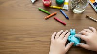 Child hands playing with blue clay elephant on wooden desk surrounded by colorful art supplies and craft materials for creative learning activities.