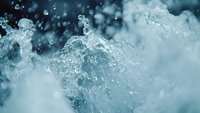 Dynamic water splash with crystalline droplets and swirling motion captured in turquoise blue tones, creating abstract patterns against blurred background.