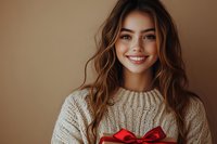 Young Asian woman with bright smile holding red gift box with bow, wearing cozy knit sweater against warm brown background. Holiday season portrait.