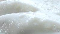 Creamy white foam texture with tiny bubbles creating soft waves and peaks, macro photography showing delicate details and light shadows for cosmetic backgrounds.