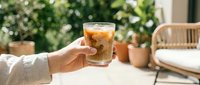 Person holding glass of iced tea or cold brew coffee outdoors on sunny patio with green plants and wooden furniture in background.