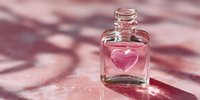 Small glass perfume bottle with pink heart-shaped design on front, casting soft shadows on pink surface in natural light.