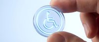 Transparent circular badge with wheelchair accessibility symbol held between human fingers against white background for inclusive design projects.
