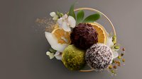 Luxury chocolate truffles with pistachio, coconut, and dark chocolate coating arranged with orange slices and white orchid flowers on golden ring decoration.