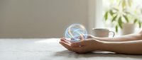 Hands holding glowing light bulb with swirling energy inside on white desk with coffee cup and plant in background representing innovation and creativity.
