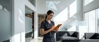 Young professional woman reading orange book in modern office lobby with natural sunlight streaming through large windows creating geometric shadows.