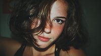 Close-up portrait of young woman with freckles and brown wavy hair against dark background, creating intimate and authentic mood.