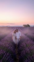 Couple walking through blooming lavender field at sunset, holding hands in romantic countryside landscape with rustic farmhouse and mountains in background.