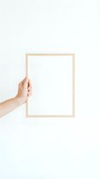 Hand holding blank wooden picture frame against white wall for mockup design and artwork display.