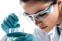 Young Asian female scientist in protective eyewear and gloves conducting laboratory research, examining liquid sample in test tube against bright background.