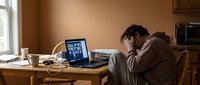 Stressed businessman holding head in hands at home office desk with laptop during video conference call experiencing work burnout and mental fatigue.