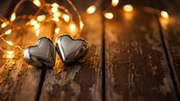 Two silver heart-shaped ornaments on rustic wooden surface with warm golden fairy lights creating romantic bokeh background for Valentine's Day celebrations.