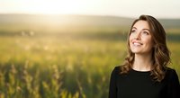 Young woman with long brown wavy hair and fair complexion gazing upward in a lush green agricultural field during golden hour sunset, wearing black clothing.