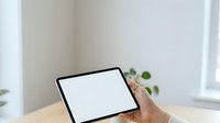 Cropped view of male hand holding tablet with blank white screen on wooden desk near window with potted plant for app mockup.