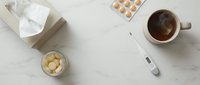 Overhead view of sick person's essentials including tissues, medicine pills, thermometer, hot tea and ginger on marble surface for cold and flu care.
