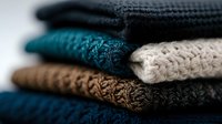 Stack of folded knitted sweaters in various colors including teal, beige, brown and navy blue showing detailed wool texture and patterns in soft focus.