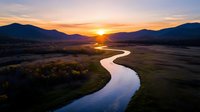 Meandering river reflects golden sunset light as it winds through mountain valley landscape, with dramatic sky and silhouetted peaks creating serene wilderness scene.